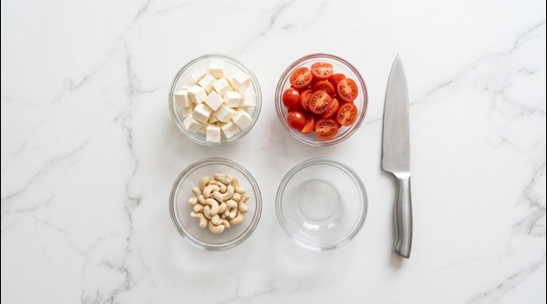 Diced paneer cubes soaking in a bowl of water next to fresh tomatoes and cashews Diced paneer cubes soaking in a bowl of water next to fresh tomatoes and cashews